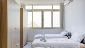 A hotel room with two beds, a wardrobe, a window with a view, and an air conditioning unit. - Seafront 3BR APT in Sliema with Private Balcony (Sliema)
