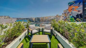 A rooftop terrace with two chairs and a table, surrounded by greenery, overlooking a cityscape and a body of water.