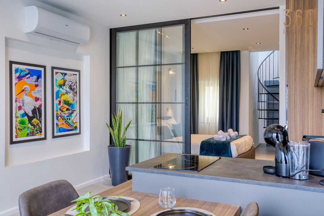 Ein modernes Hotelzimmer mit einer großen Glasdoor, die auf einen Balkon führt, einem Essbereich mit Tisch und Stühlen sowie zwei gerahmten Kunstwerken an der Wand.
