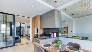 Una cocina moderna con una isla central, un comedor con una mesa larga dispuesta para seis personas, y una escalera que lleva a un nivel superior.