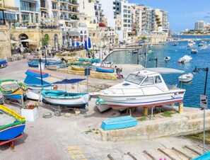 A marina with boats docked, a stone quay, and buildings in the background.