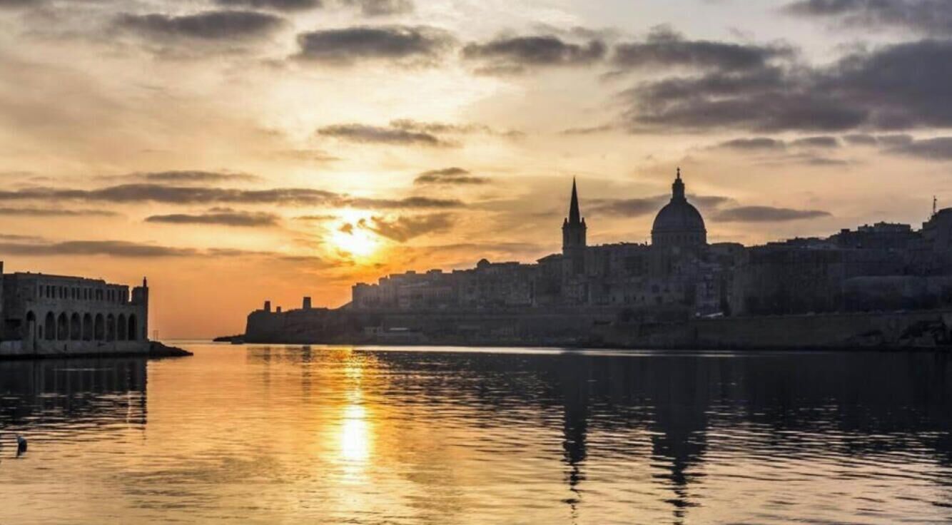 A cityscape with a prominent church and a dome, reflecting on calm water during sunset.