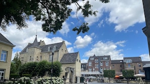 Un edificio storico con una torre imponente, un cortile lastricato e vegetazione circostante.