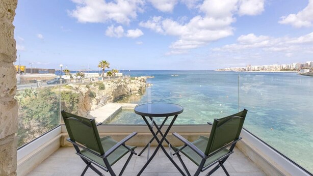 A balcony with a table and chairs overlooking a coastal area with clear blue water and a rocky shoreline. - Unique studio with Breathtaking views in St Pauls (San Pawl il-Baħar)
