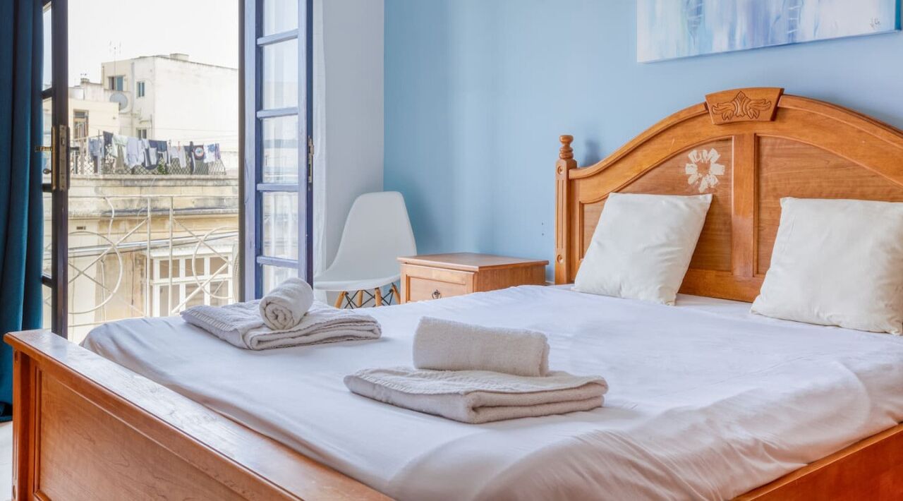 A neatly made bed with white linens and towels, a wooden headboard with a floral design, a small bedside table, and a view of buildings through a window.