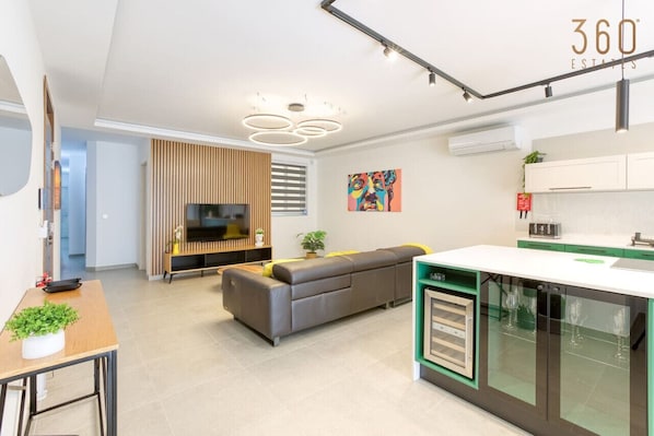 Eine moderne Küche mit einer zentralen Kochinsel, ein Essbereich mit Tisch und Stühlen sowie ein Wohnzimmer mit Sofa und Fernseher.