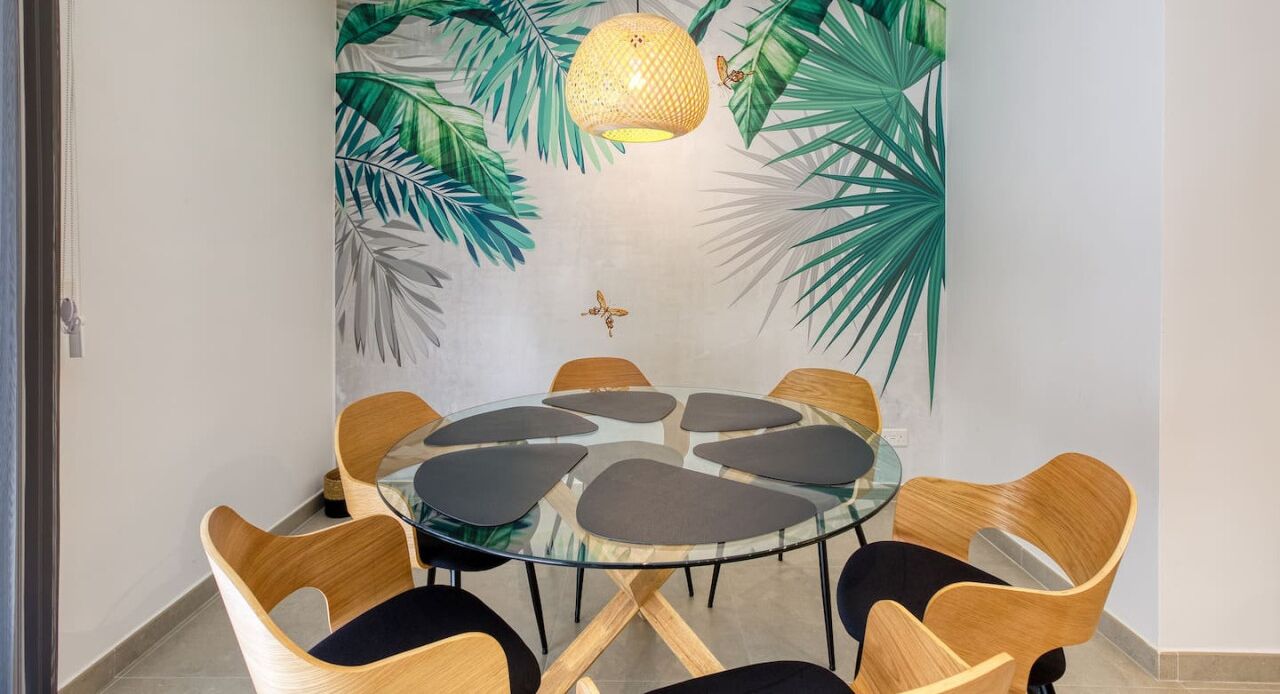 A round glass table with six chairs, a hanging light fixture, and a wall mural with tropical leaves.
