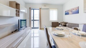 Un salon moderne et bien éclairé, avec une table à manger pour quatre personnes, un téléviseur à écran plat et une grande fenêtre donnant sur la ville.