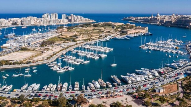 Une vue aérienne d’une marina où sont amarrés de nombreux bateaux, entourée d’un paysage urbain côtier avec des bâtiments et un ciel bleu dégagé.