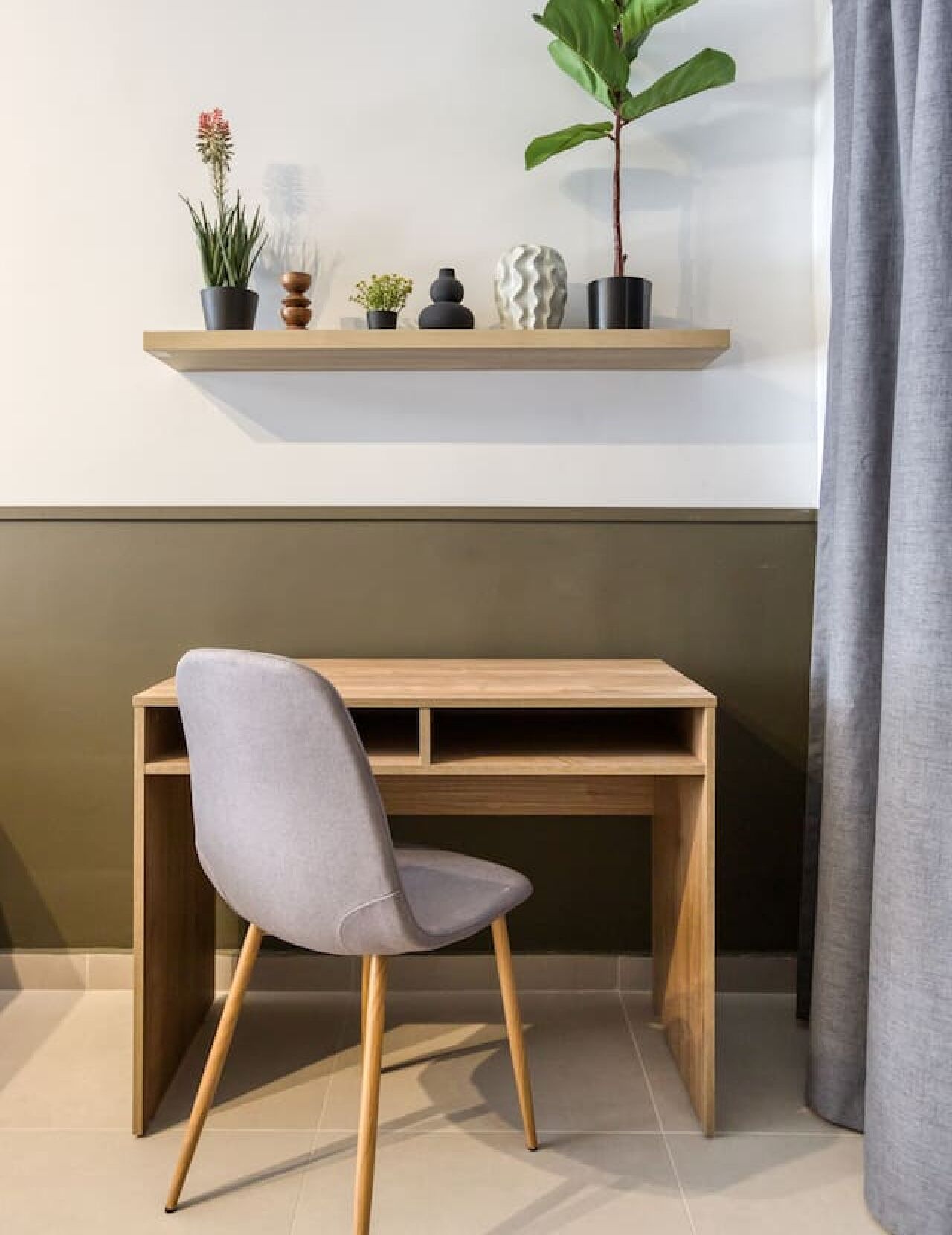 Un escritorio moderno con una silla gris, una estantería de madera encima con varios objetos decorativos, y una cortina gris a la derecha.