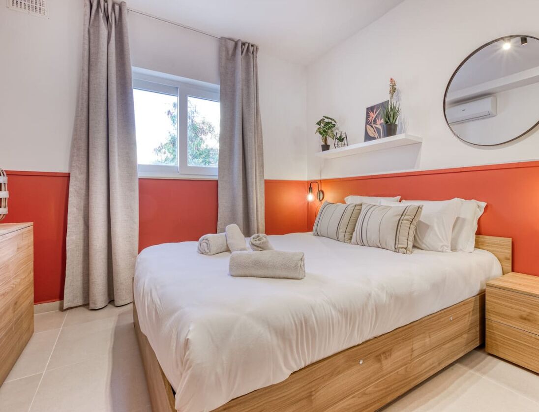 A bedroom with a bed, bedside tables, a window with curtains, a wall-mounted mirror, and a shelf with plants.