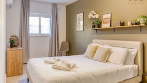 A modern bedroom with a large bed, a wooden nightstand, a chair, and a window with curtains. There are wall-mounted shelves with decorative items and a sign that reads "360 ESTATES". - A lovely, cozy fully equipped 2BR home in Pieta (Tal-Pietà)