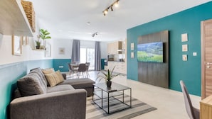 A modern living room with a sofa, coffee table, and TV. There is a kitchen in the background with cabinets and a dining area. - A lovely, cozy fully equipped 2BR home in Pieta (Tal-Pietà)