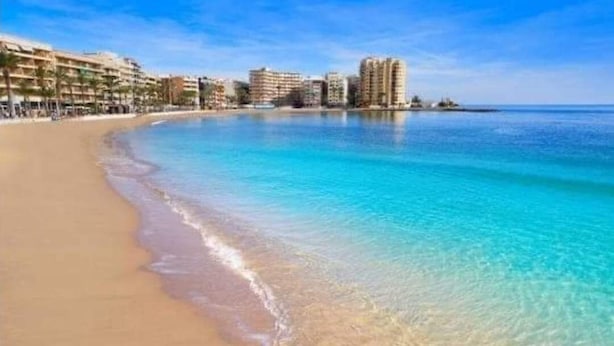 Una playa con agua turquesa cristalina, una orilla arenosa y edificios al fondo.