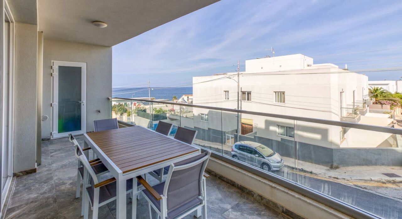 A balcony with a table and chairs, a glass railing, and a view of the sea and buildings.