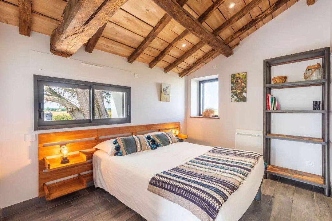 Ein Schlafzimmer mit einem großen Bett, einem Holzkopfstück, Nachttischen, einem Regal und einem Fenster mit Blick auf Bäume.