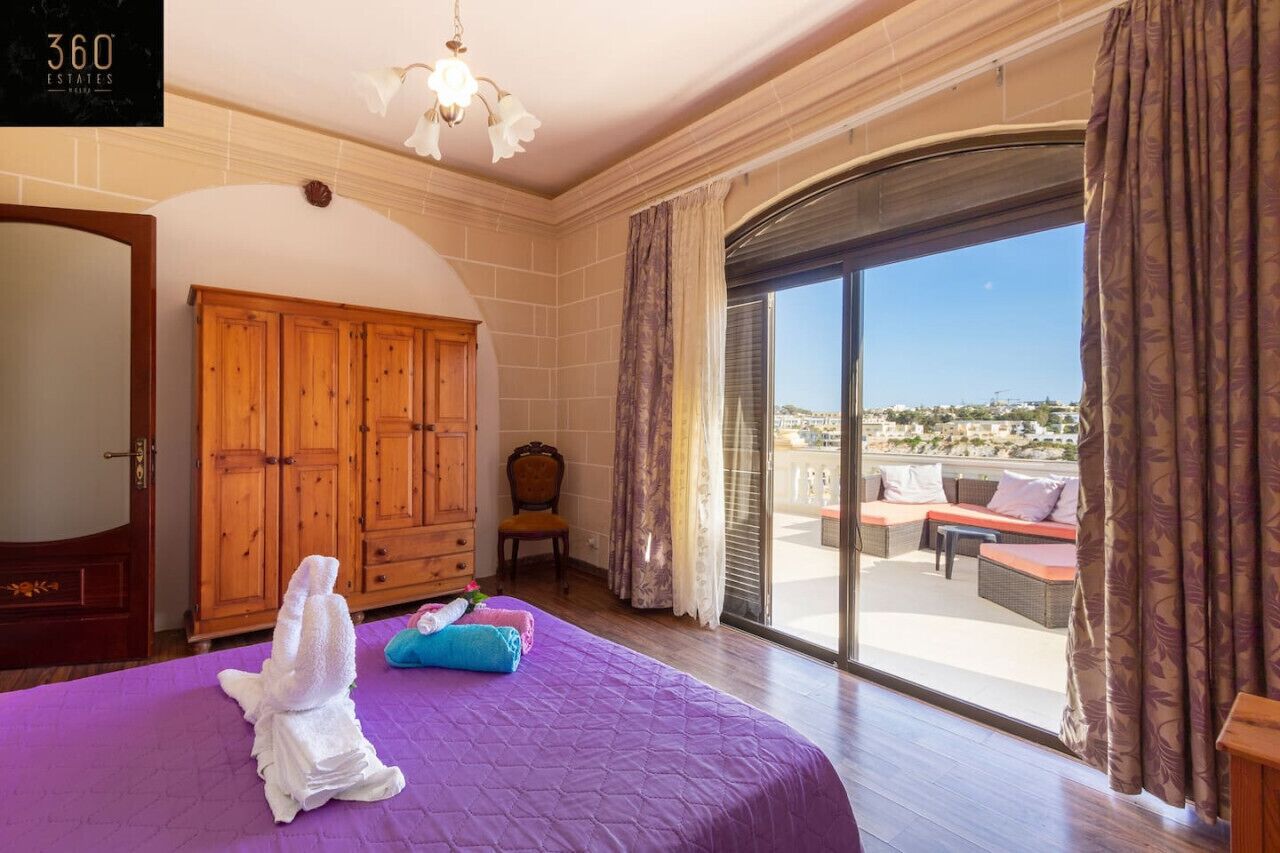 A hotel room with a large bed, a wooden wardrobe, a chair, and a balcony with outdoor furniture.