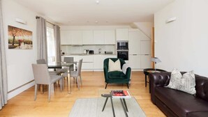 A modern apartment with a dining area, a green armchair, a leather sofa, and a kitchen in the background.