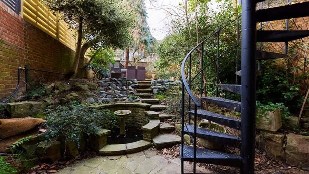 A stone pathway leading to a circular water feature, surrounded by rocks and greenery, with a spiral staircase on the right.