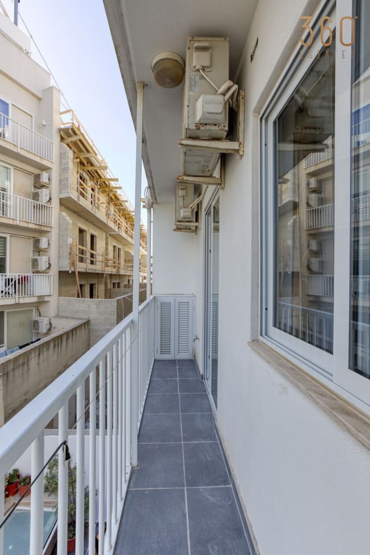 Een smal balkon met betegelde vloer, witte leuningen en een glazen deur die naar een kamer leidt. Airconditioningunits zijn aan de muur bevestigd.