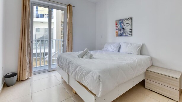 Ein Schlafzimmer mit einem Bett, einem Nachttisch, einem Gemälde und Blick auf ein Gebäude durch eine Schiebetür.