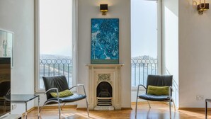A spacious living room with a fireplace, two chairs, a large window, and a city view.