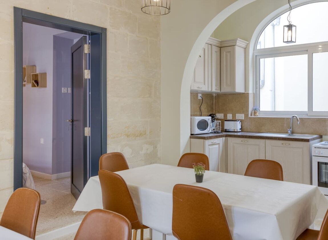 A dining area with a table set for six, a kitchen with white cabinets and a window, and a door leading to another room.