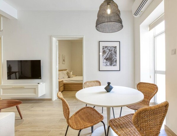A modern living room with a round white table, wicker chairs, a flat-screen TV, and a large window.