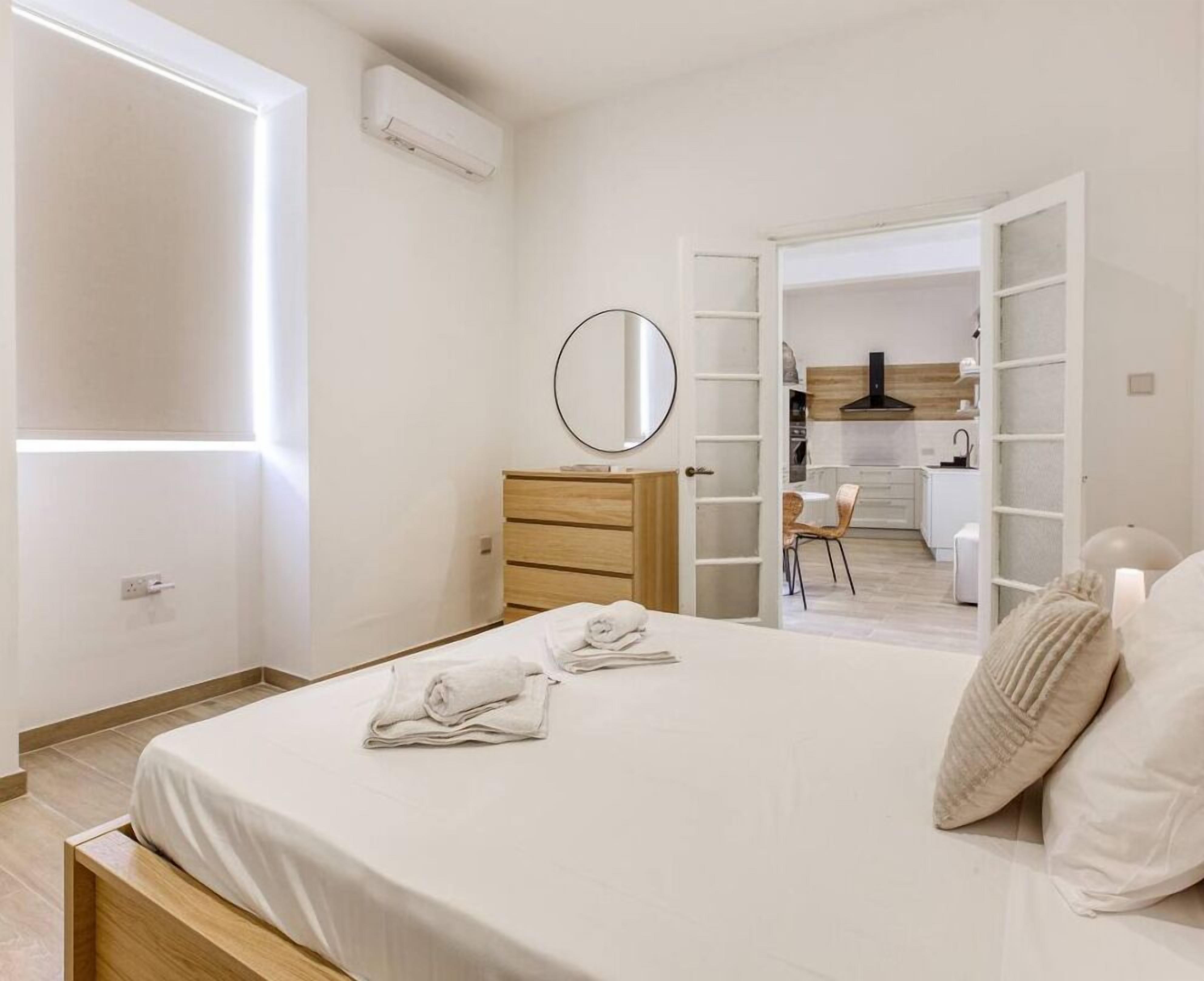 Ein Schlafzimmer mit Bett, Nachttisch, Spiegel und Blick in einen Küchenbereich.