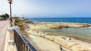 A coastal walkway with a rocky beach, clear blue sea, and a few people sunbathing.