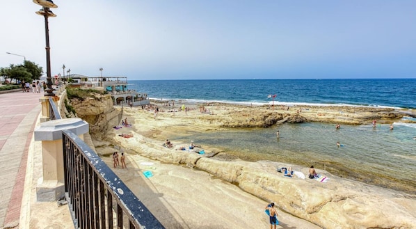 Un camino costero con una playa rocosa, mar azul cristalino y algunas personas tomando el sol.