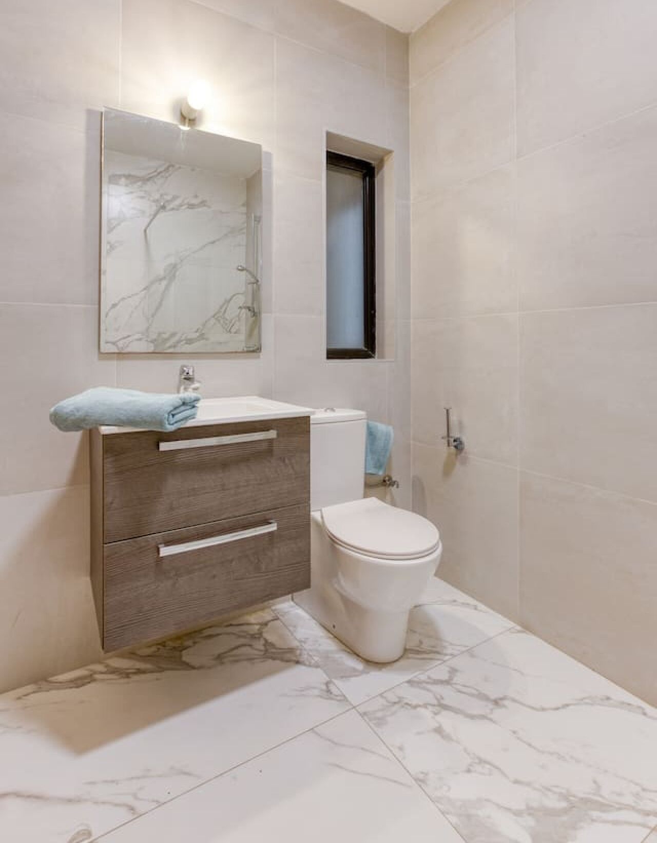 Um banheiro com piso de mármore, vaso sanitário branco, toucador de madeira com espelho e uma toalha no balcão.