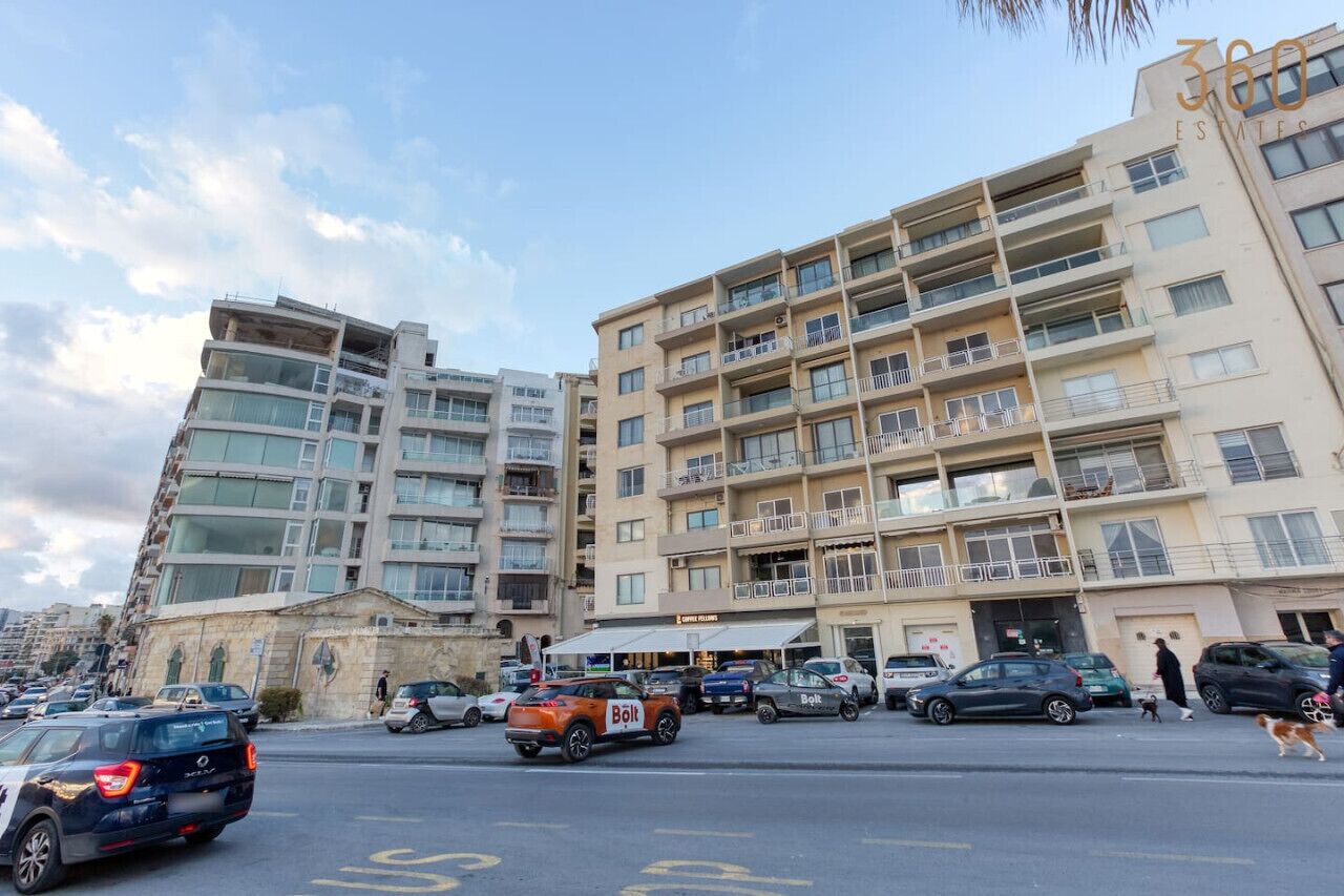 Un edificio a più piani con balconi, un'area di parcheggio e una strada con auto e pedoni.