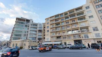 Un edificio a più piani con balconi, un'area di parcheggio e una strada con auto e pedoni.