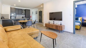 A modern living room with a sofa, a dining table with chairs, a TV mounted on the wall, and a kitchen area in the background. - Spacious 3BR home with private Balconies in Mosta (Il-Mosta)