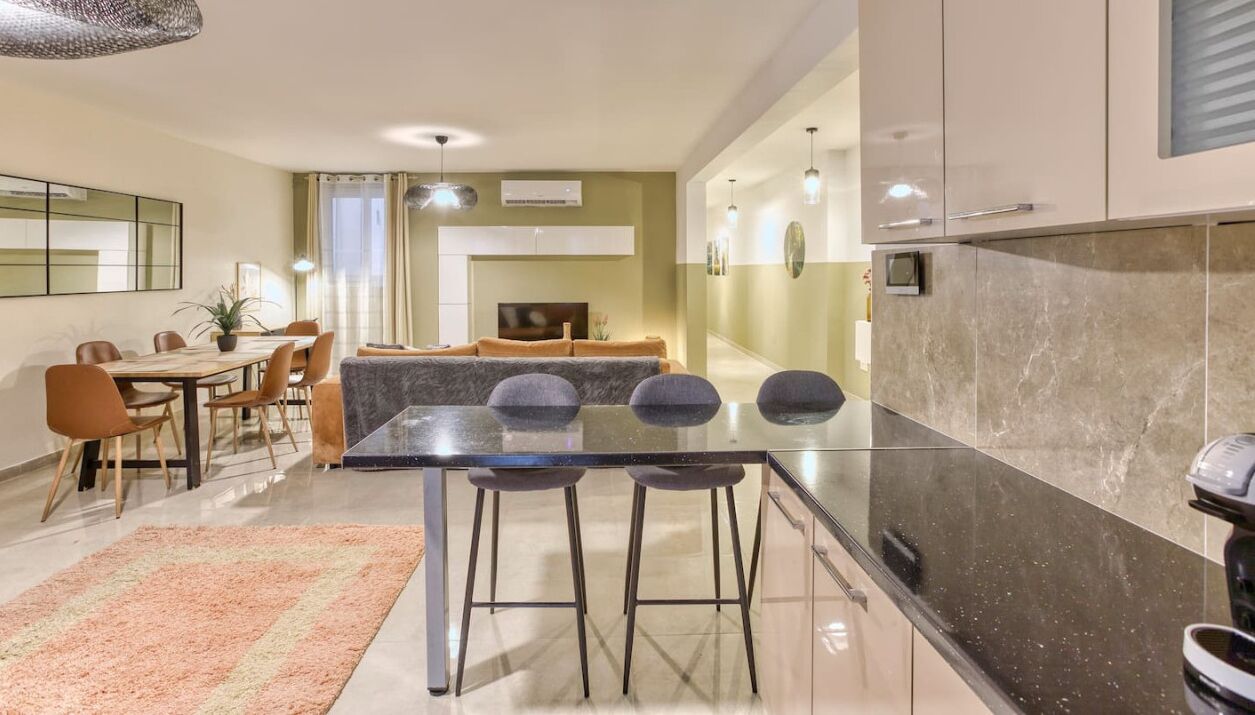 A modern kitchen with a marble countertop, a dining area with a glass table, and a living room with a sofa and TV.