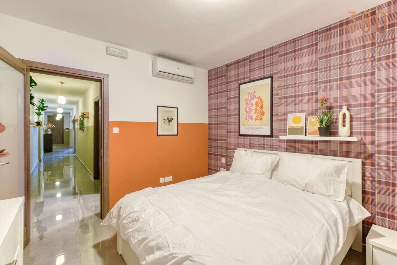 A hotel room with a bed, a mirror, an air conditioning unit, a hallway, and a wall with a plaid pattern.