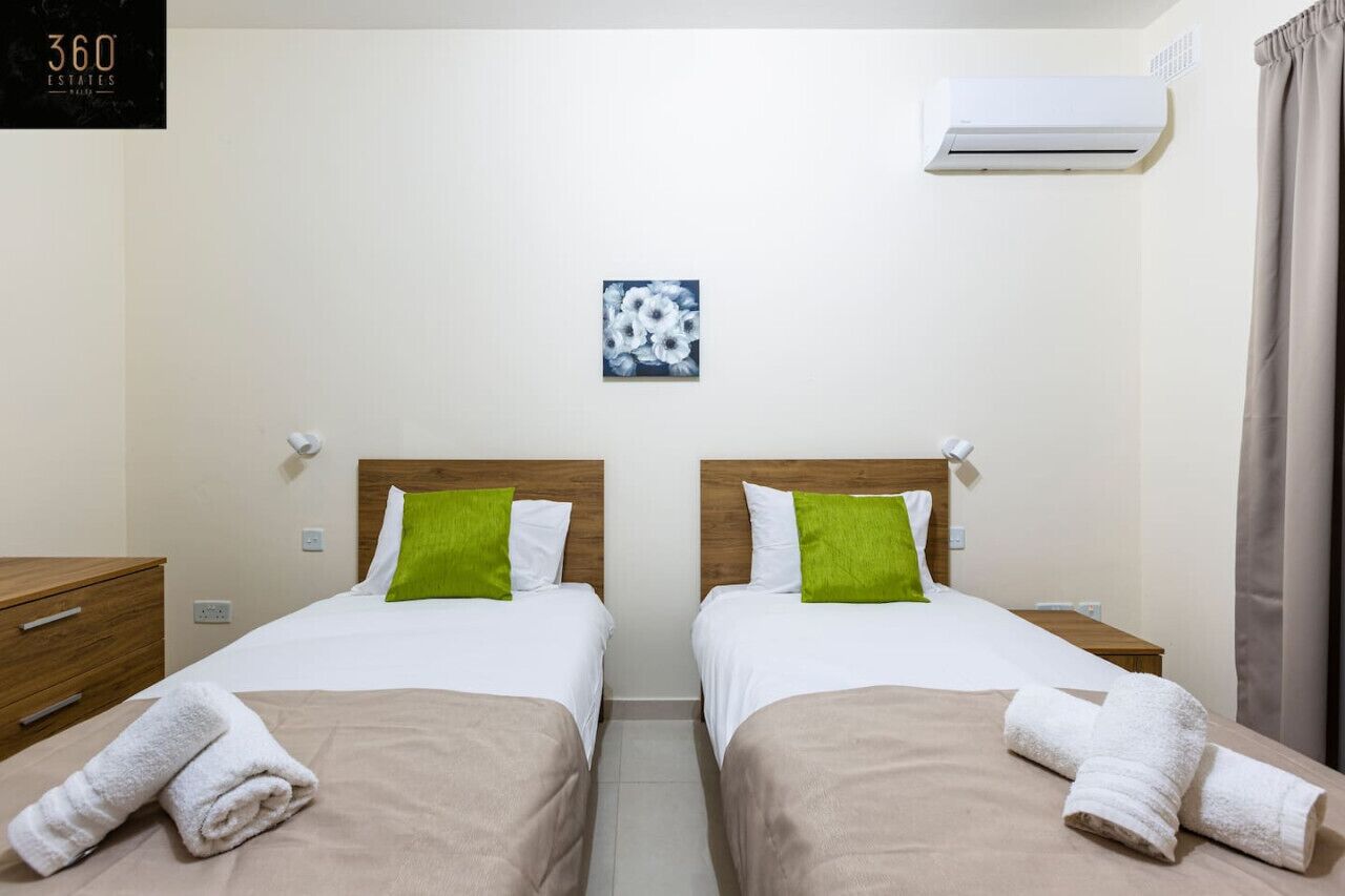 A hotel room with two beds, each with a wooden headboard, white bedding, and a green pillow. There is a small artwork on the wall, an air conditioning unit above the beds, and a wooden nightstand with a lamp.