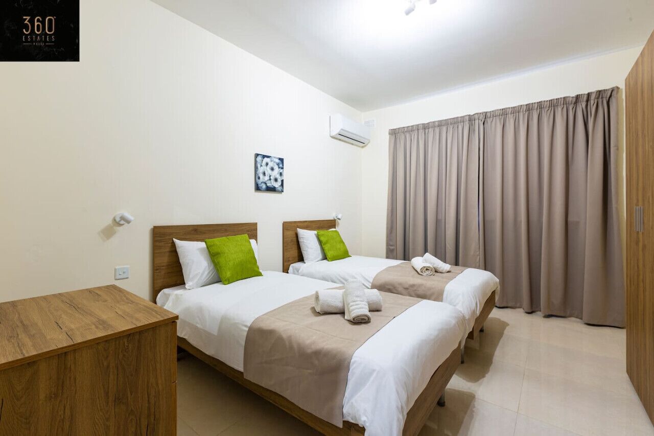 Une chambre d’hôtel avec deux lits, une table de chevet en bois, un climatiseur fixé au mur et une grande fenêtre avec des rideaux.