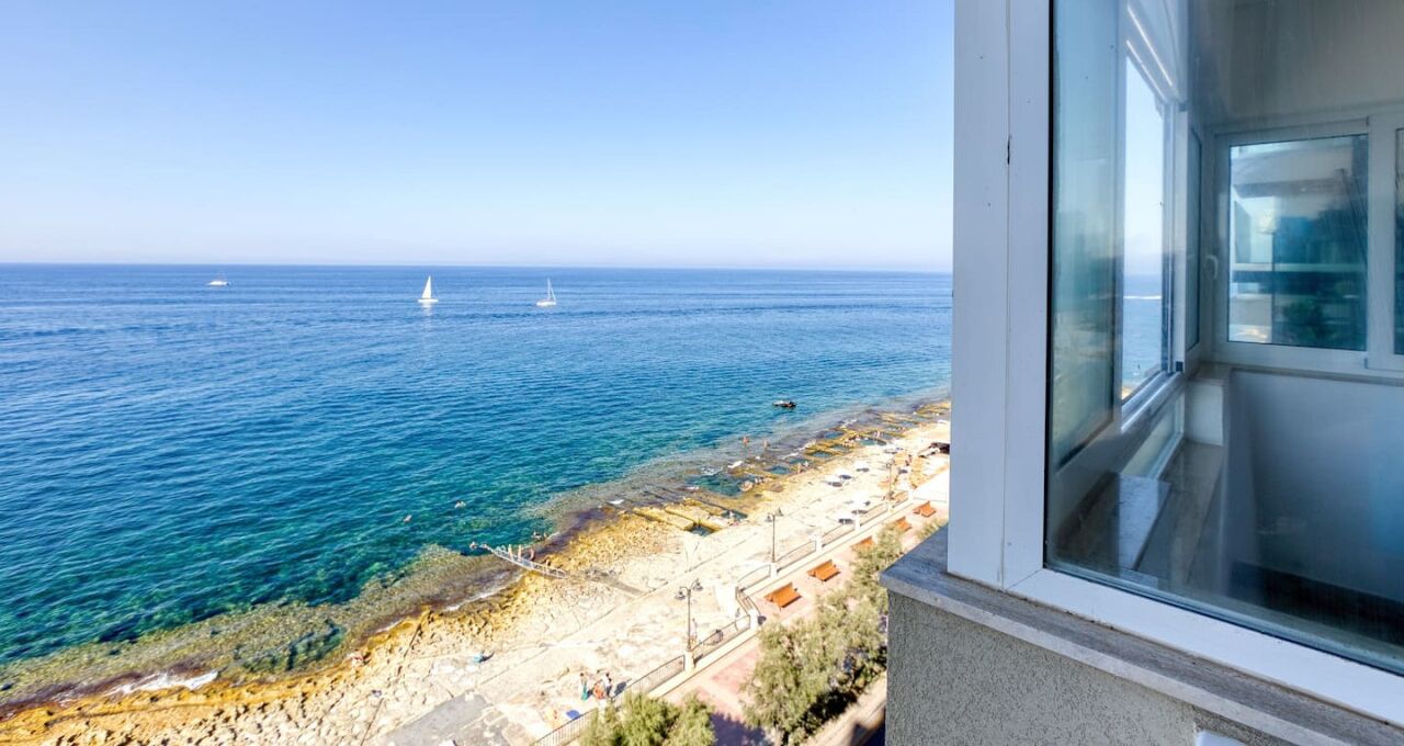 A coastal view from a window overlooking a beach with clear blue waters, a few boats, and a sandy shore with some structures.