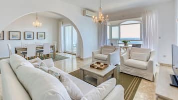 Un ampio soggiorno con un divano spazioso, due poltrone, un tavolino da caffè e un'area pranzo con tavolo e sedie. Sono presenti due lampadari, una televisione e vista mare attraverso ampie finestre.