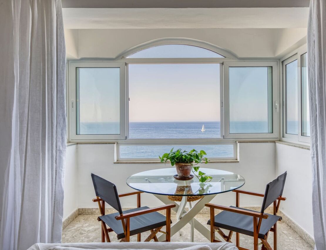A balcony with a glass table and chairs, a potted plant, and a view of the sea.