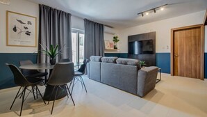 A modern living room with a grey sofa, a dining table with chairs, a flat-screen TV mounted on the wall, and a wooden door. - Modern 2BR Apartment with Private Balcony in Pieta (Tal-Pietà)