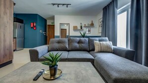 A modern living room with a grey sectional sofa, a coffee table with a plant, a remote control, and a wall-mounted fire extinguisher. - Modern 2BR Apartment with Private Balcony in Pieta (Tal-Pietà)