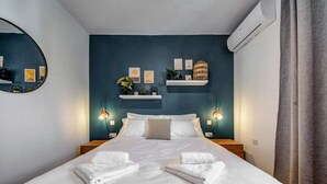 Un letto rifatto con lenzuola e asciugamani bianchi, un condizionatore d'aria a parete e mensole decorative con piante e quadri.