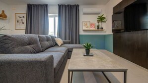 A modern living room with a grey sofa, a coffee table, and a TV. There are curtains, a plant, and framed pictures on the walls. - Modern 2BR home with Balcony & Sofa bed in Pieta (Tal-Pietà)