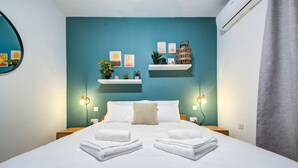 A neatly made bed with white linens and two folded towels in the center. There are two bedside tables with lamps, a wall-mounted shelf with decorative items, and a large woven basket on the shelf. - Modern 2BR home with Balcony & Sofa bed in Pieta (Tal-Pietà)