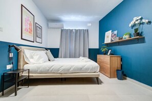 A bedroom with a bed, bedside table, and a nightstand. There is a window with curtains, a wall-mounted air conditioner, and a shelf with plants and pictures.
