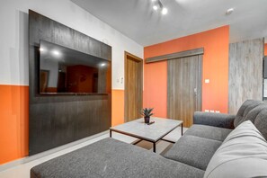 A modern living room with a grey sofa, a small table with a plant, and a large flat-screen TV mounted on a dark wall.