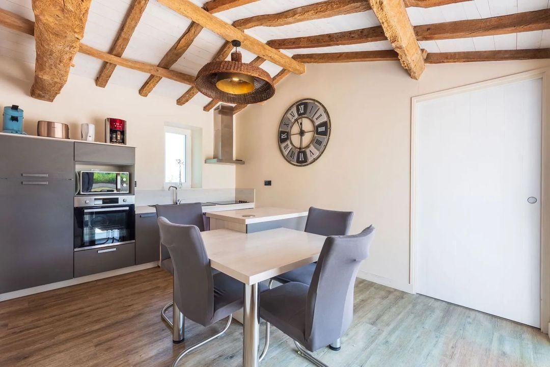 A modern kitchen with a dining area, wooden ceiling beams, a wall clock, and a white door.